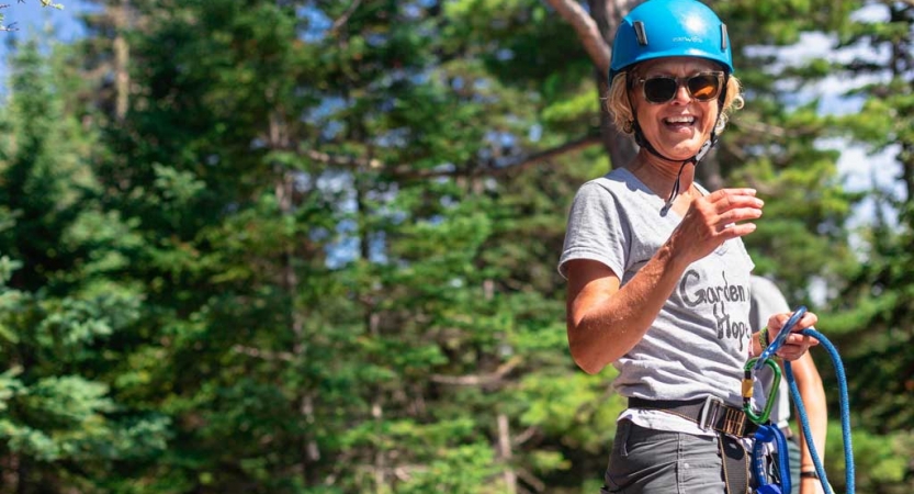 A person wearing safety gear smiles at the camera. There are green trees behind them.
